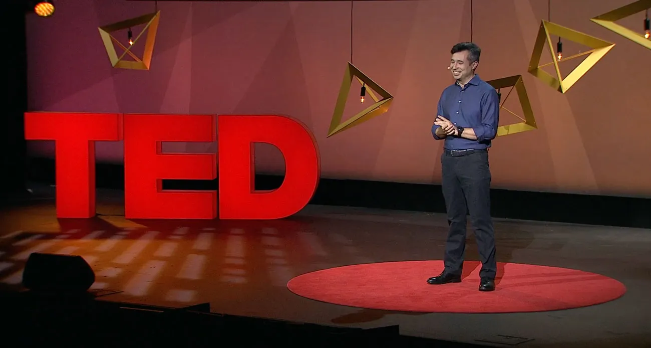 A speaker on the red circle of a TED Institute stage, the red TED letters behind him and gold wire-frame lanterns hanging overhead